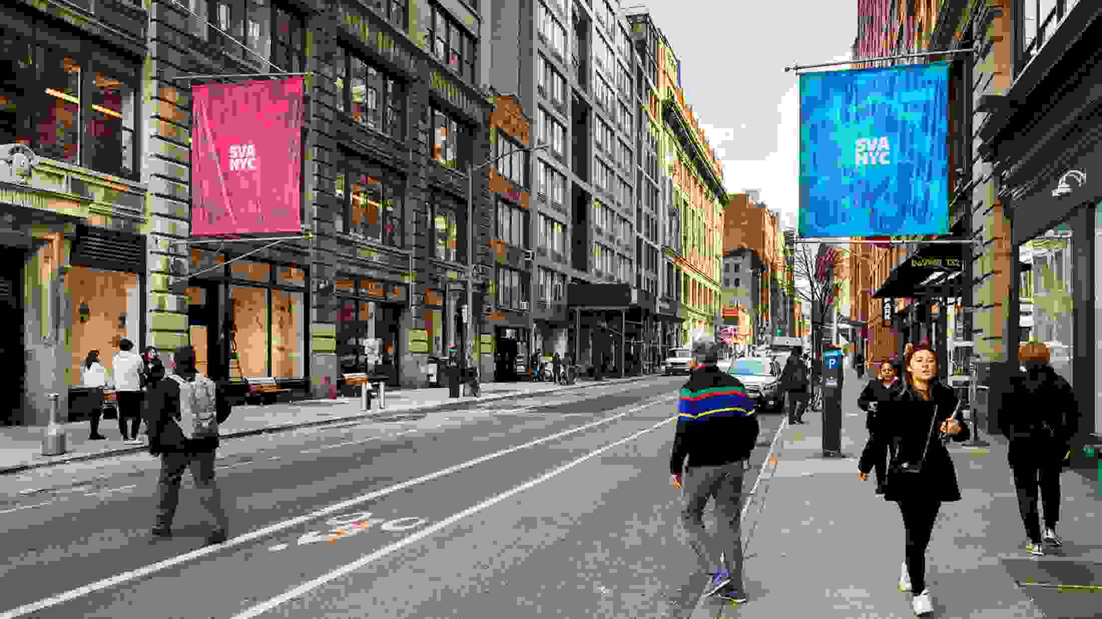 New York City street with pedestrians walking on both sides of the sidewalk as well as crossing the road; two large institutional flags are visible, the left being a red flag, flower outline and the words “SVA NYC,” the left being a blue flag, flower outline and the words “SVA NYC”