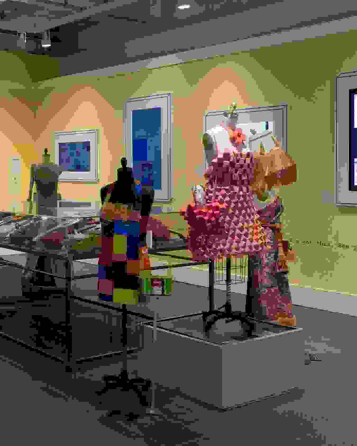 Wide view of gallery two at the Gramercy location. A large plexi vitrine on metal legs can be seen in the middle of the room, filled with painted and adorned ukuleles. Mannequins surround the vitrine and posters and monitors line the walls.