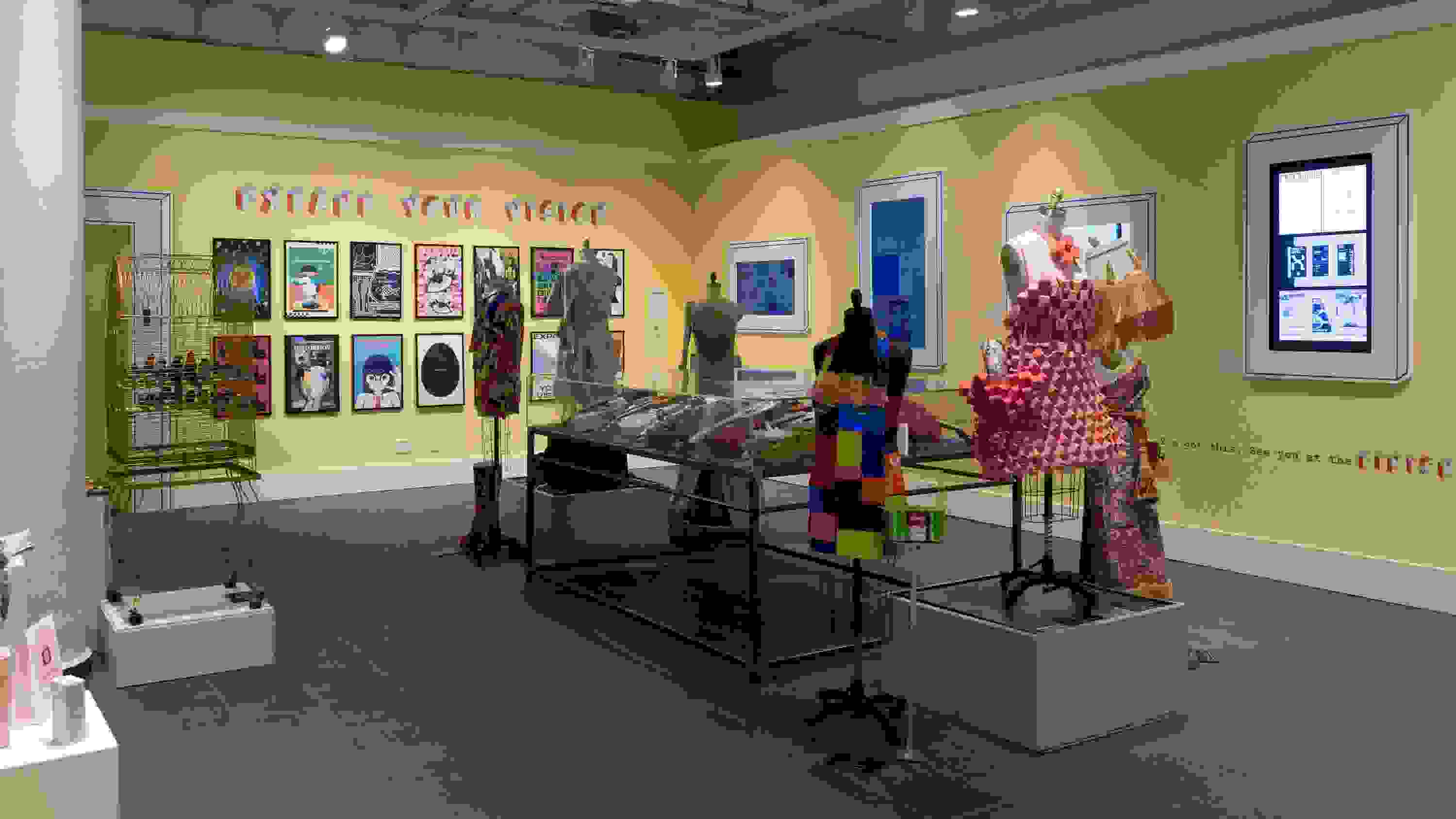 Wide view of gallery two at the Gramercy location. A large plexi vitrine on metal legs can be seen in the middle of the room, filled with painted and adorned ukuleles. Mannequins surround the vitrine and posters and monitors line the walls.