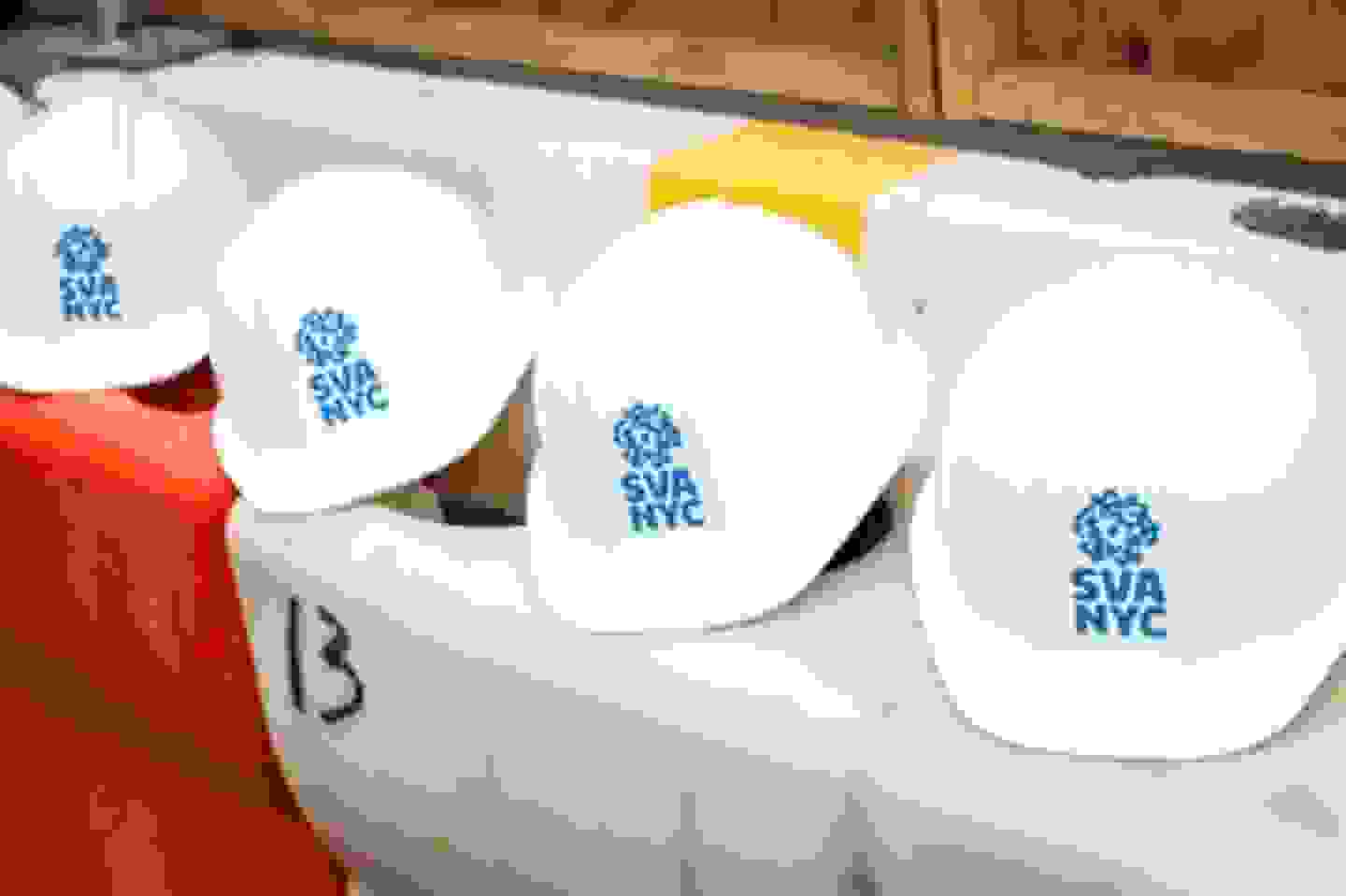 Four SVA NYC branded hard hats sit atop orange and white construction barriers
