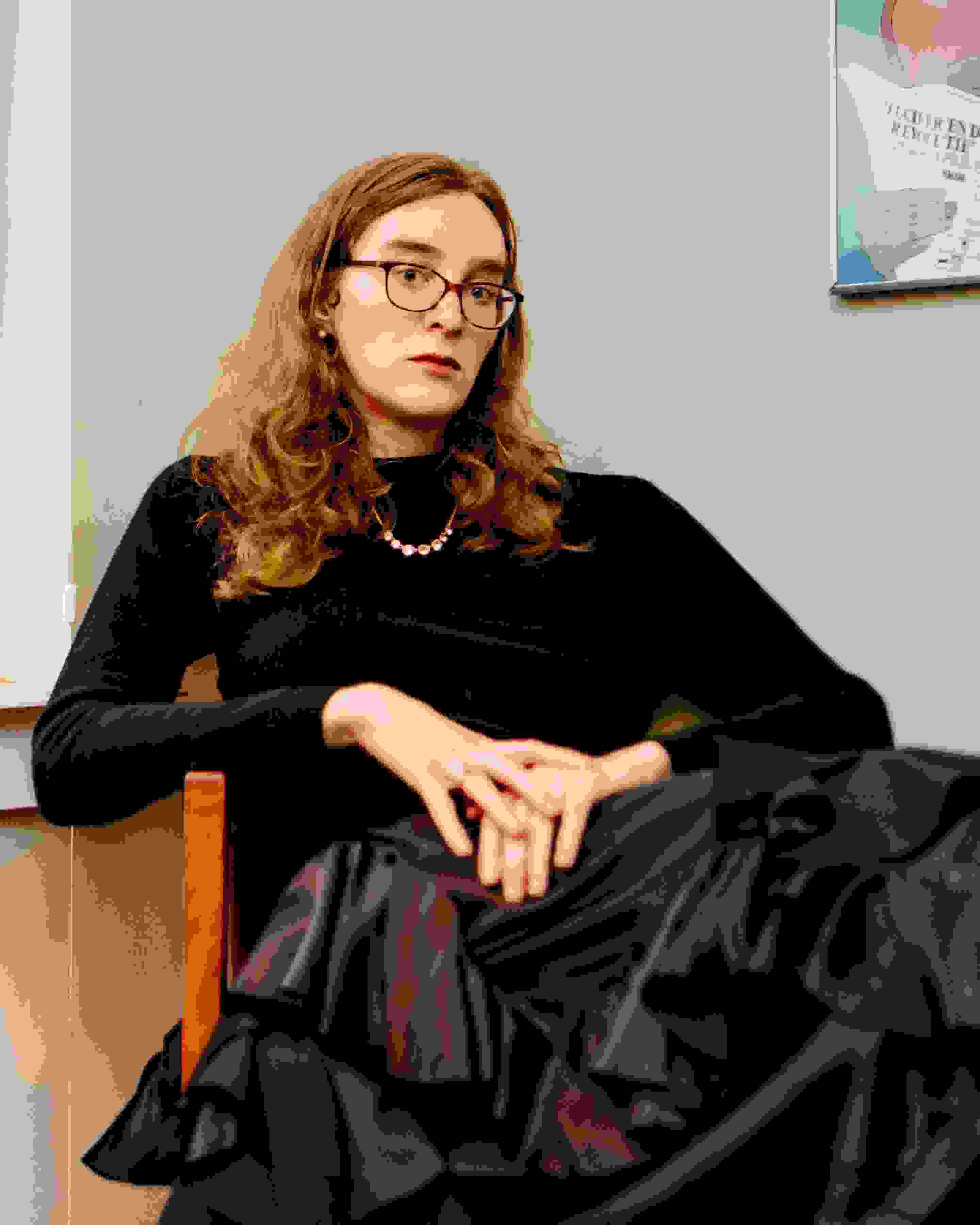 Photo portrait of the writer Grace Byron, sitting in a chair in a black turtleneck with her hands placed on top of one another. Grace wears glasses and looks directly at the lens.