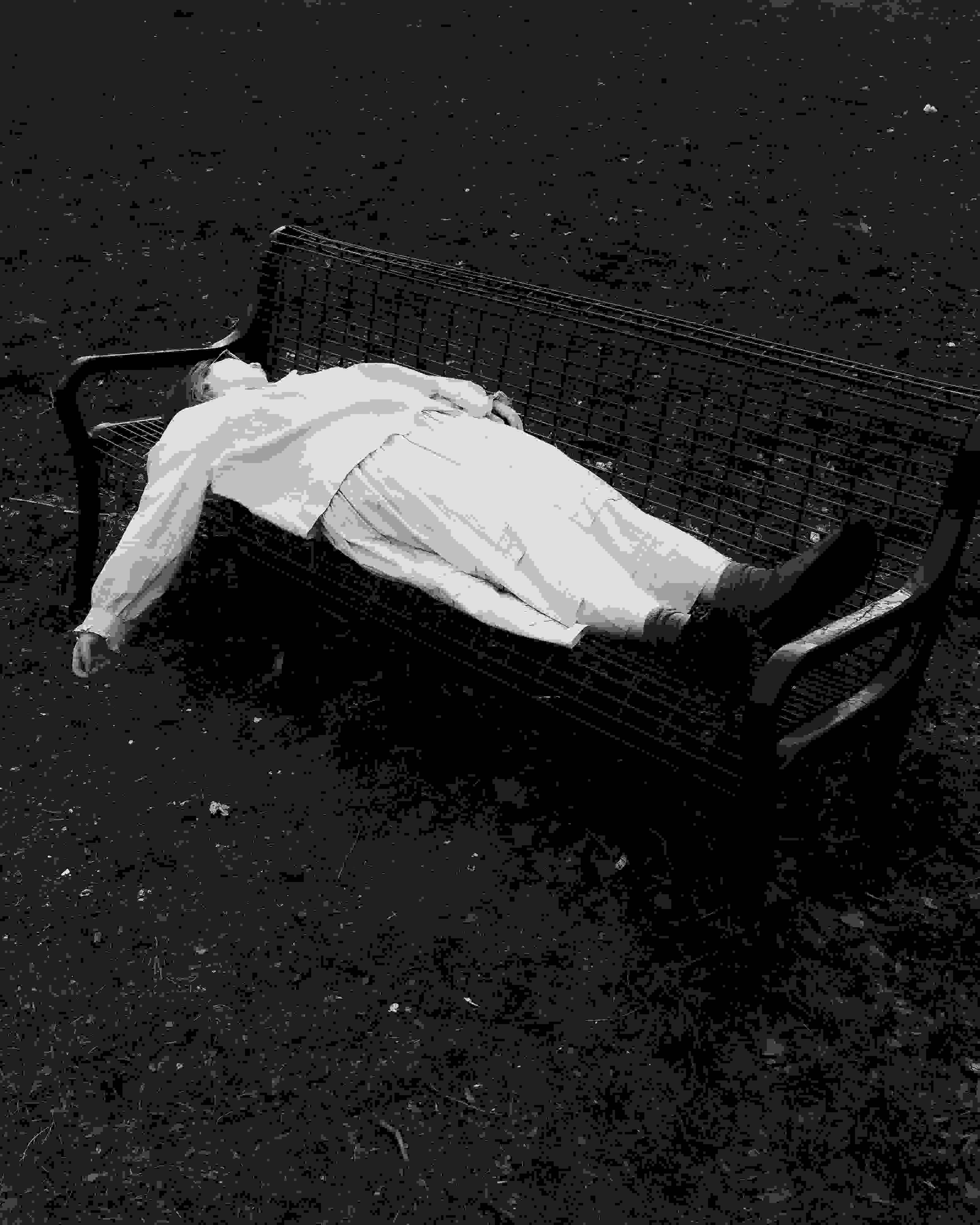 A black and white photograph of a girl lying on a bench, her hand hanging loosely, face obscured. The bench and her body are surrounded by chaotic bundles of grass.
