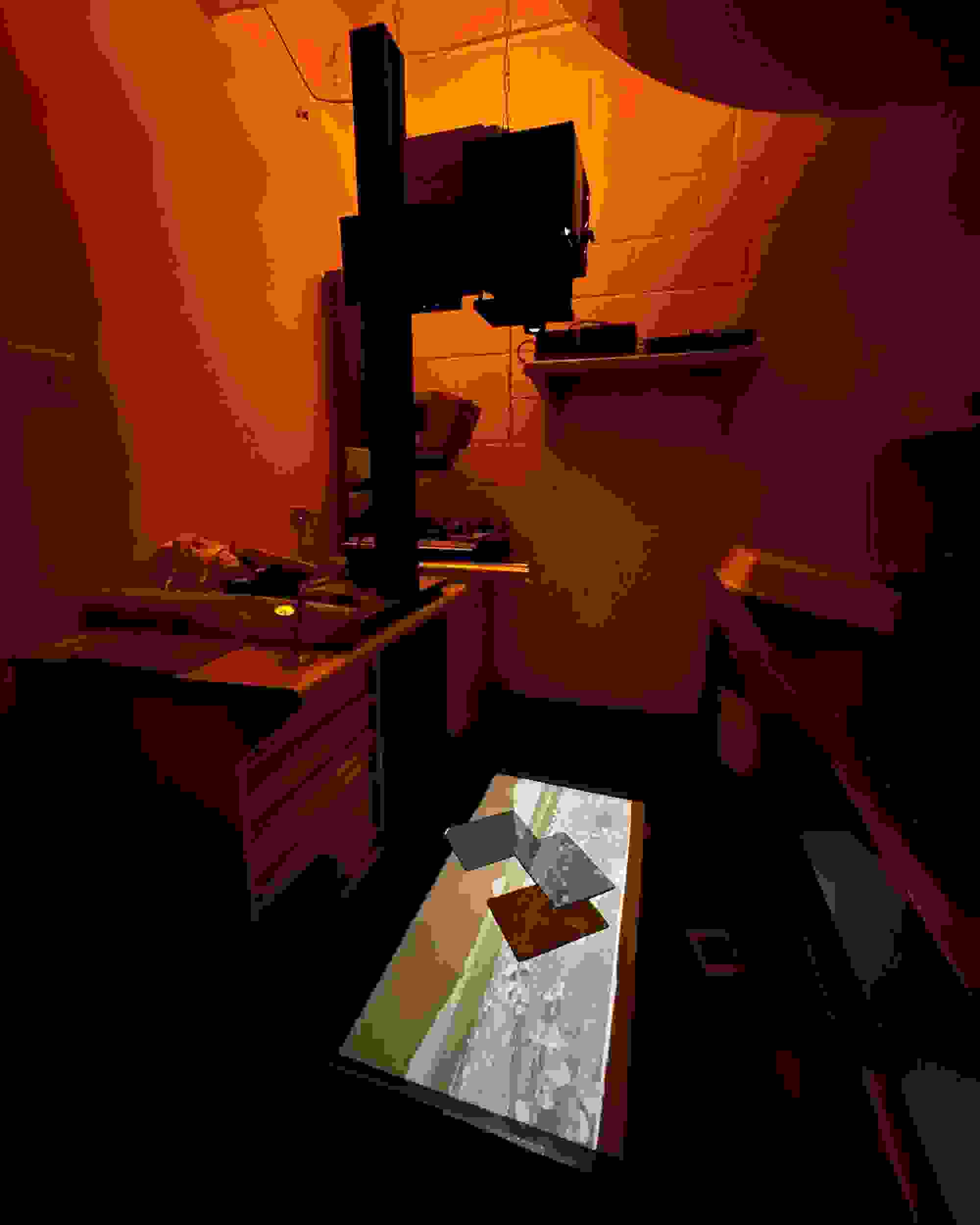 Dimly lit darkroom with an orange hue, featuring an enlarger projecting onto a surface. The emerging image creates an atmosphere of focus and mystery.