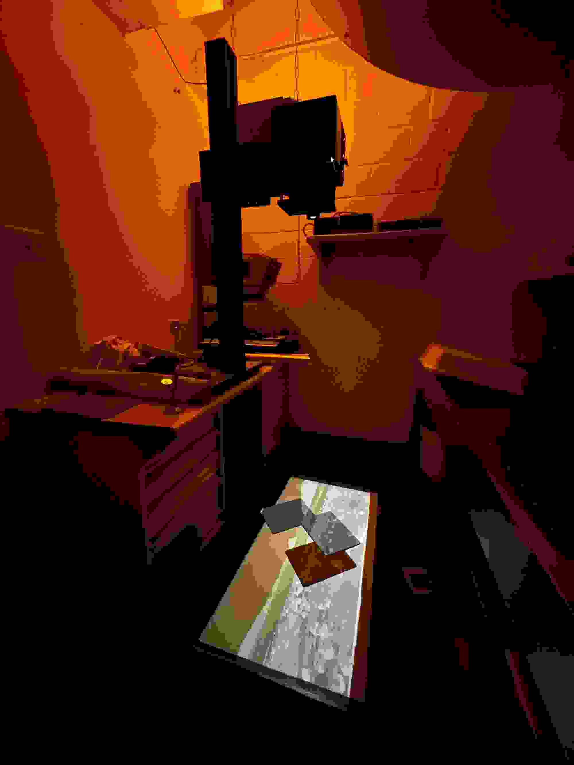 Dimly lit darkroom with an orange hue, featuring an enlarger projecting onto a surface. The emerging image creates an atmosphere of focus and mystery.