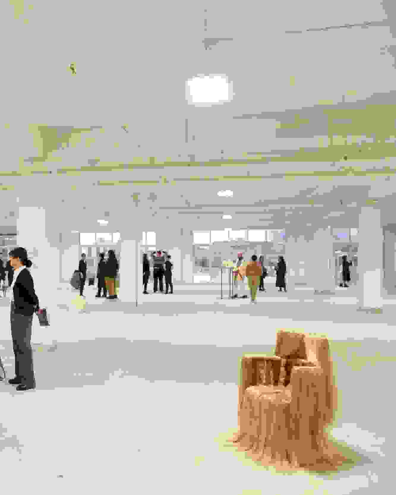 Wide view of an exhibition opening in a loft space with floor to ceiling windows. Many people mill about; there is a chair adorned with hair on display as well as art hanging on the wall.