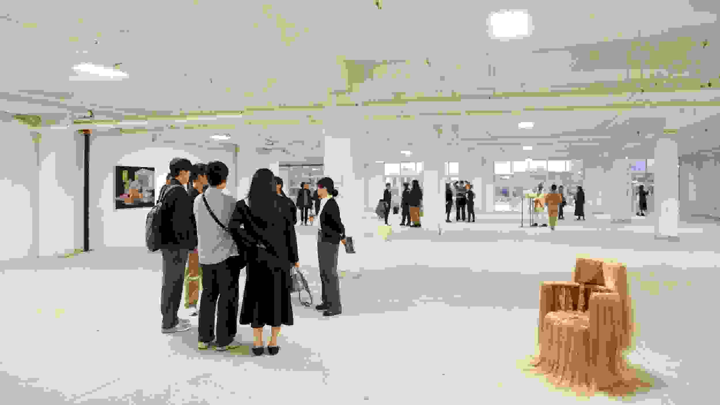 Wide view of an exhibition opening in a loft space with floor to ceiling windows. Many people mill about; there is a chair adorned with hair on display as well as art hanging on the wall.