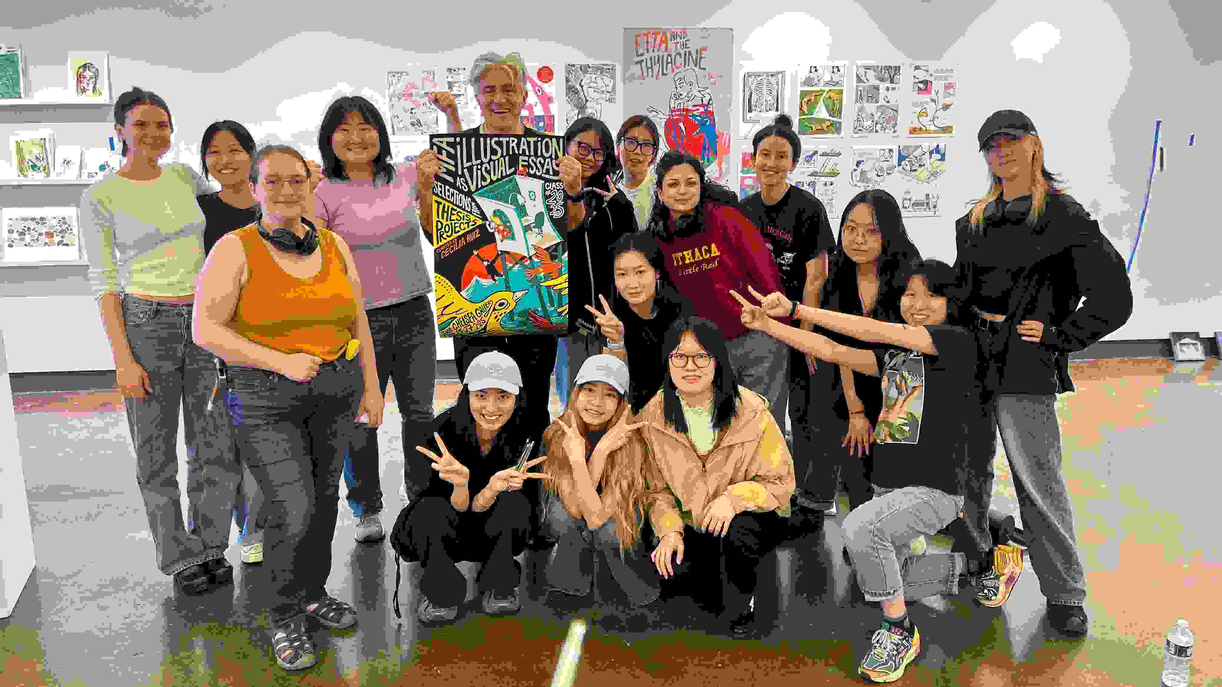 Students pose for a picture in a gallery with an instructor, who is showing off a poster design that they may have all worked on.