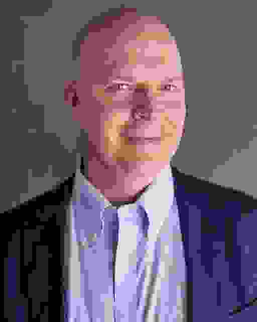 A man in a sky blue collared shirt and darker blue suit jacket smiles into