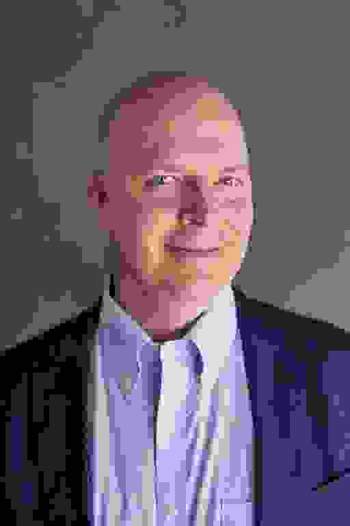 A man in a sky blue collared shirt and darker blue suit jacket smiles into