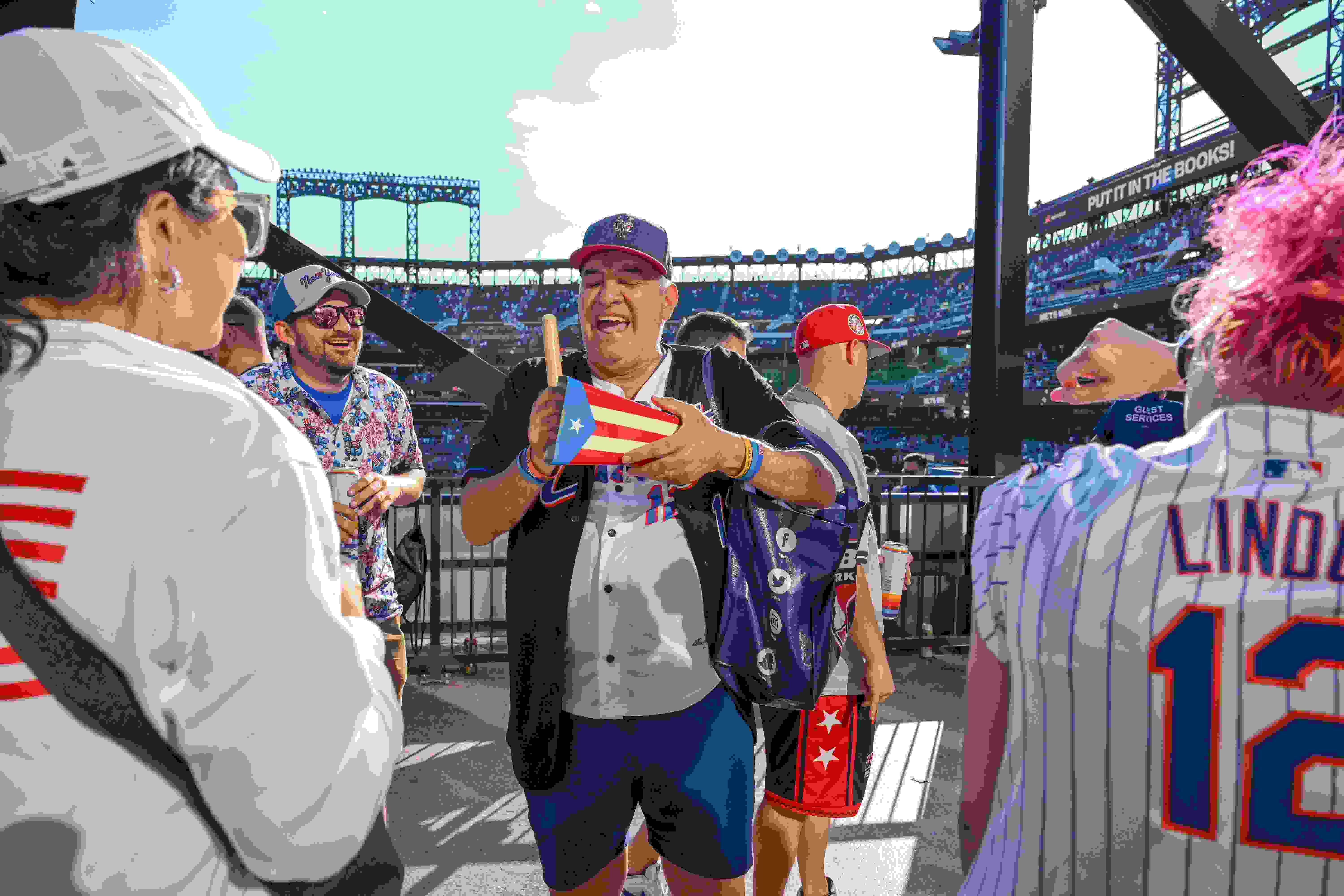 A photograph of New York Mets fans having a joyous interaction at Citi Field. The central figure is holding a cowbell painted like the Puerto Rican flag.