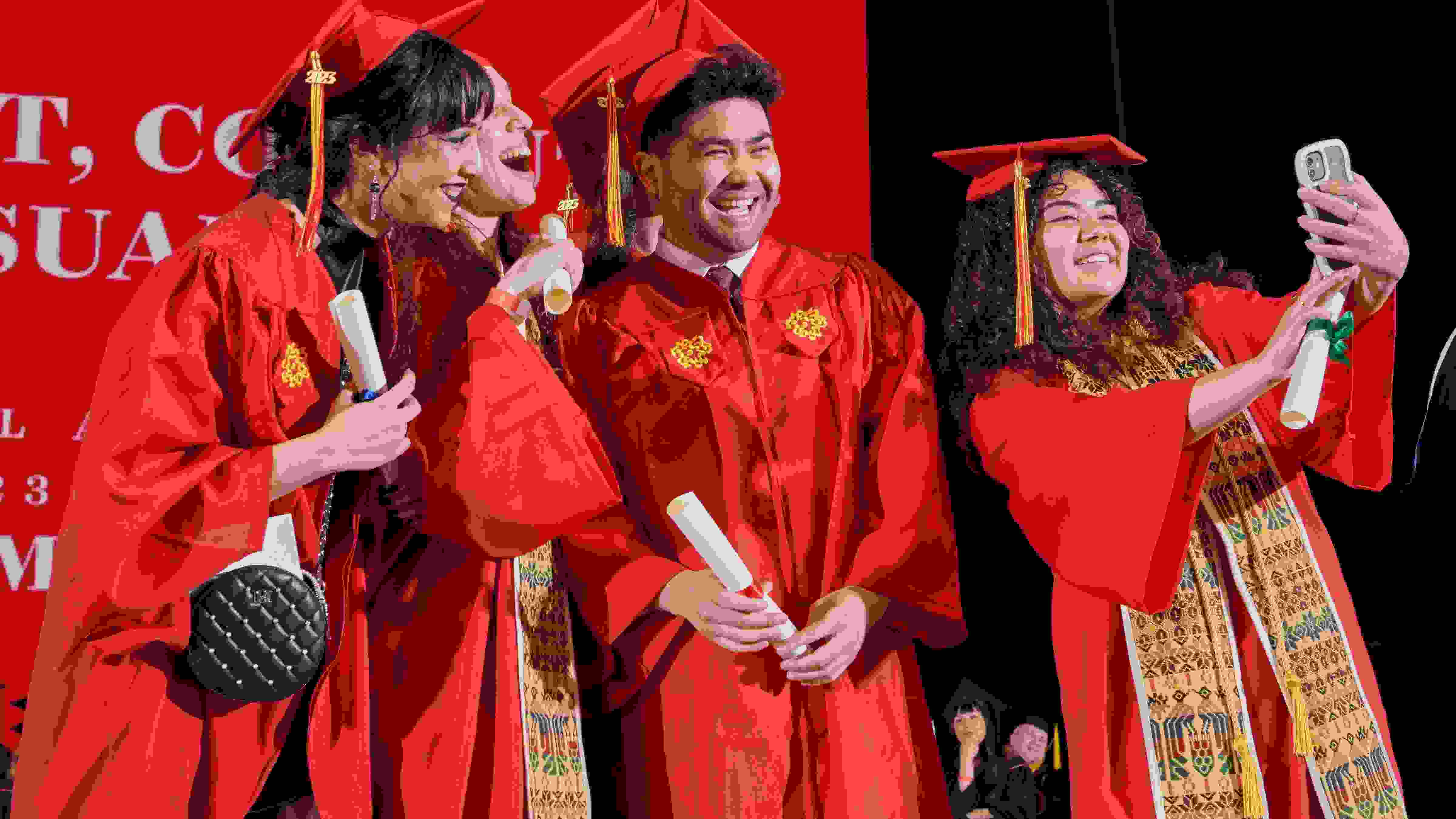 SVA graduating students in red caps and gowns, some holding flowers, smiling and posing for photos inside Radio City Music Hall in New York.