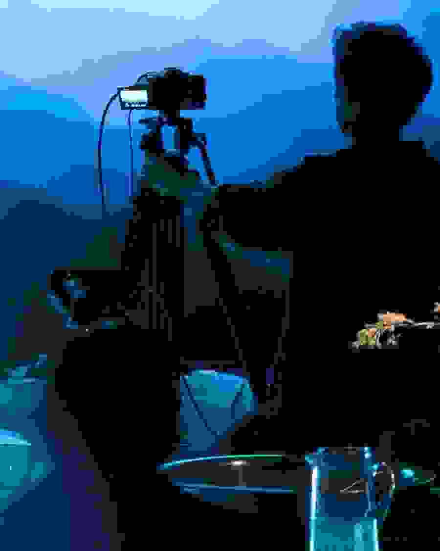  Image of a person pointing a recording camera on a tripod towards a wall with a projected image of what looks like a mountain range.