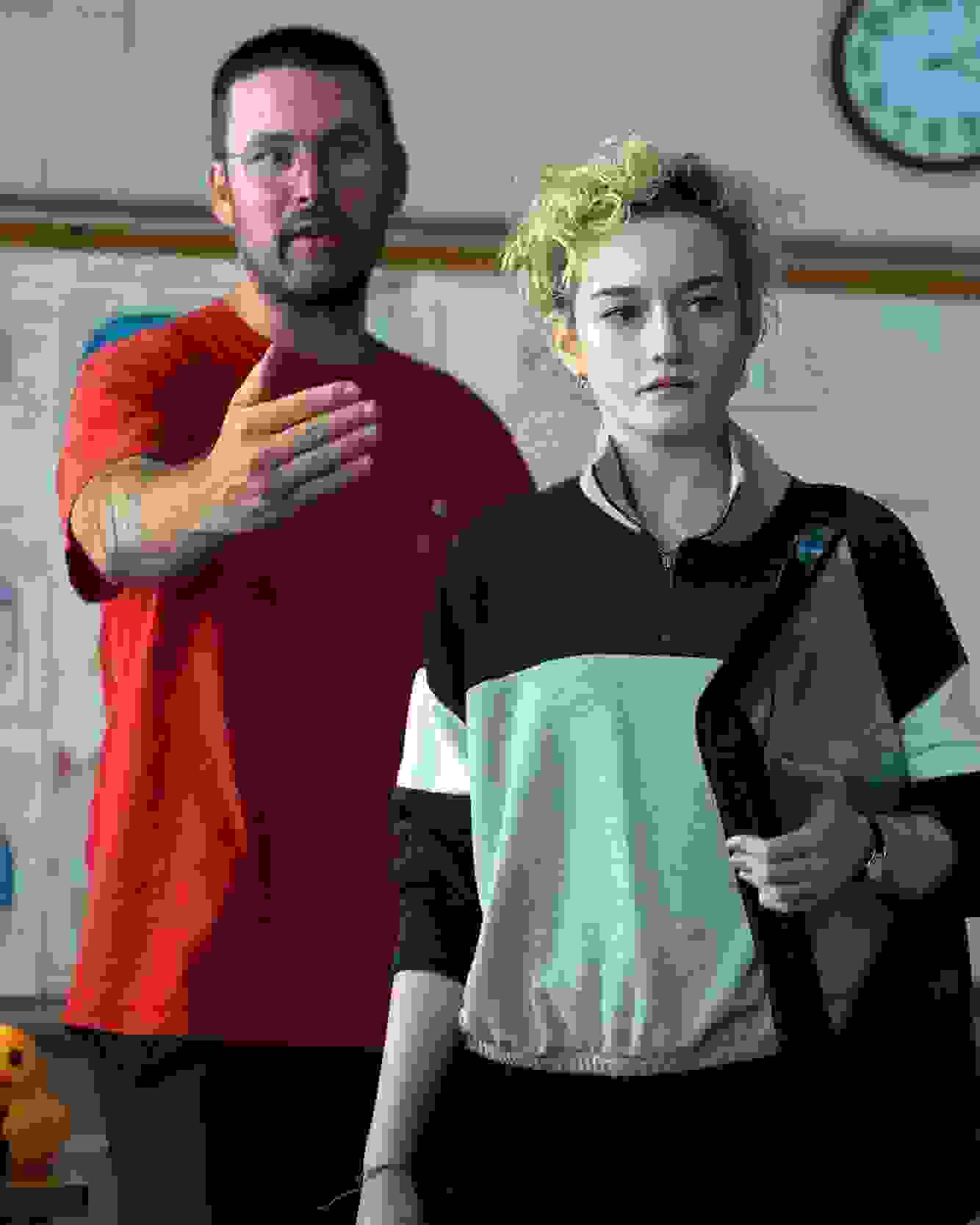 A person in a red shirt gestures in a classroom, appearing explanatory, while another with short curly hair listens intently. A whiteboard and clock are in the background.