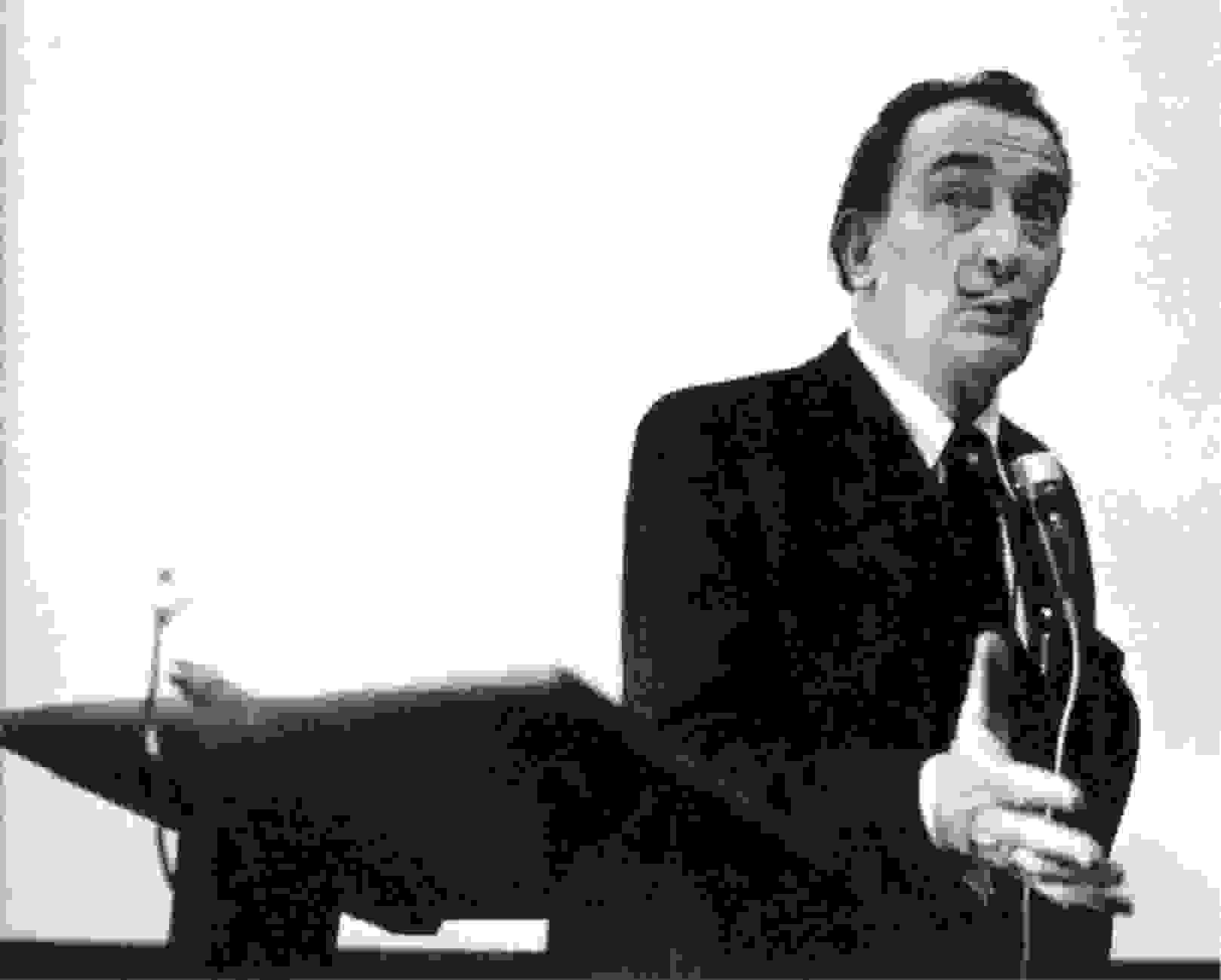 A black and white photograph of Salvador Dalí ostensibly giving a speech at a dais, sporting his signature mustache