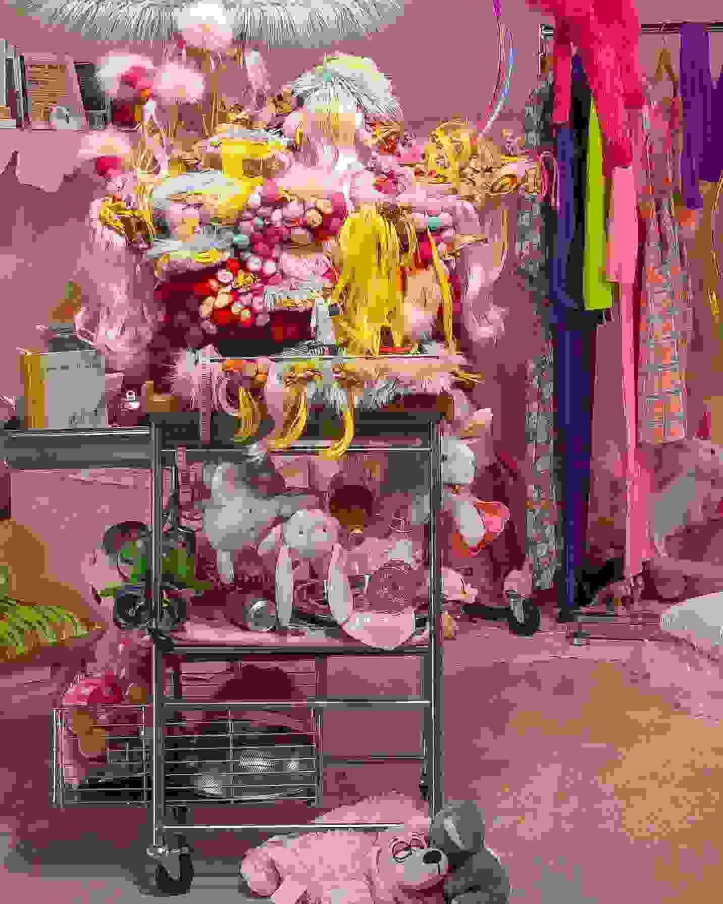 An assemblage of a clothing rack with trendy garments, artfully arranged hula hoops which intersect one another and hang down from the ceiling in a mobile-like fashion. Under the mobile, a sculpture-like assemble of stuffed animals and toys stacked intricately. behind all the items is a baby pink painted wall, which also contains a shelf displaying framed art and ephemera