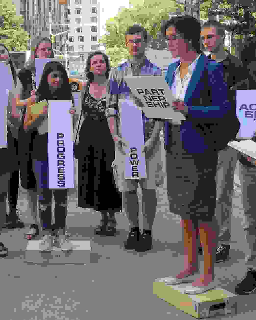 Group of students standing on platforms outdoors, holding placards marked with various design concepts