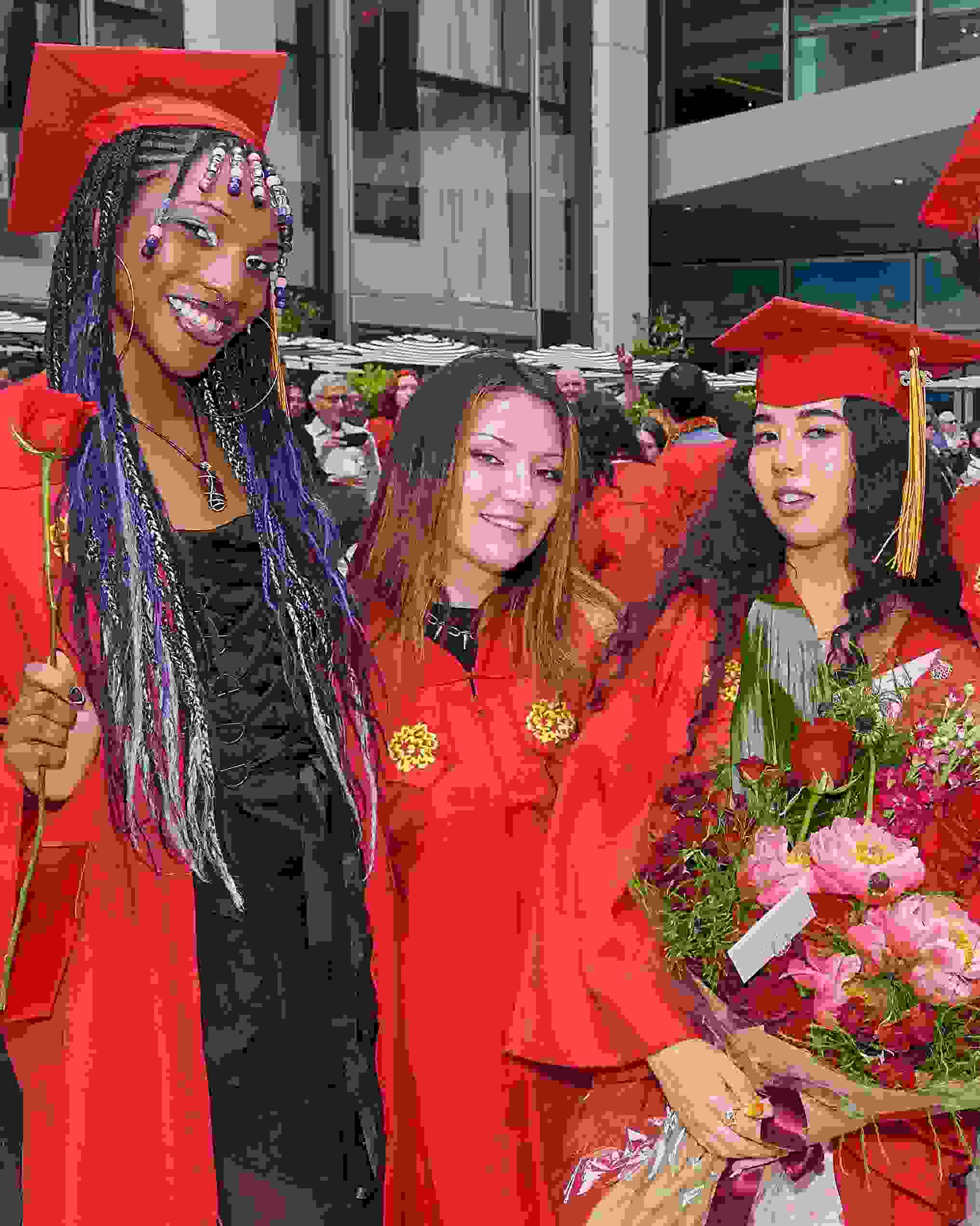 Three graduates wearing red commencement regalia pose for a photo; the right-most person is holding a large bouquet of pink and red flowers.