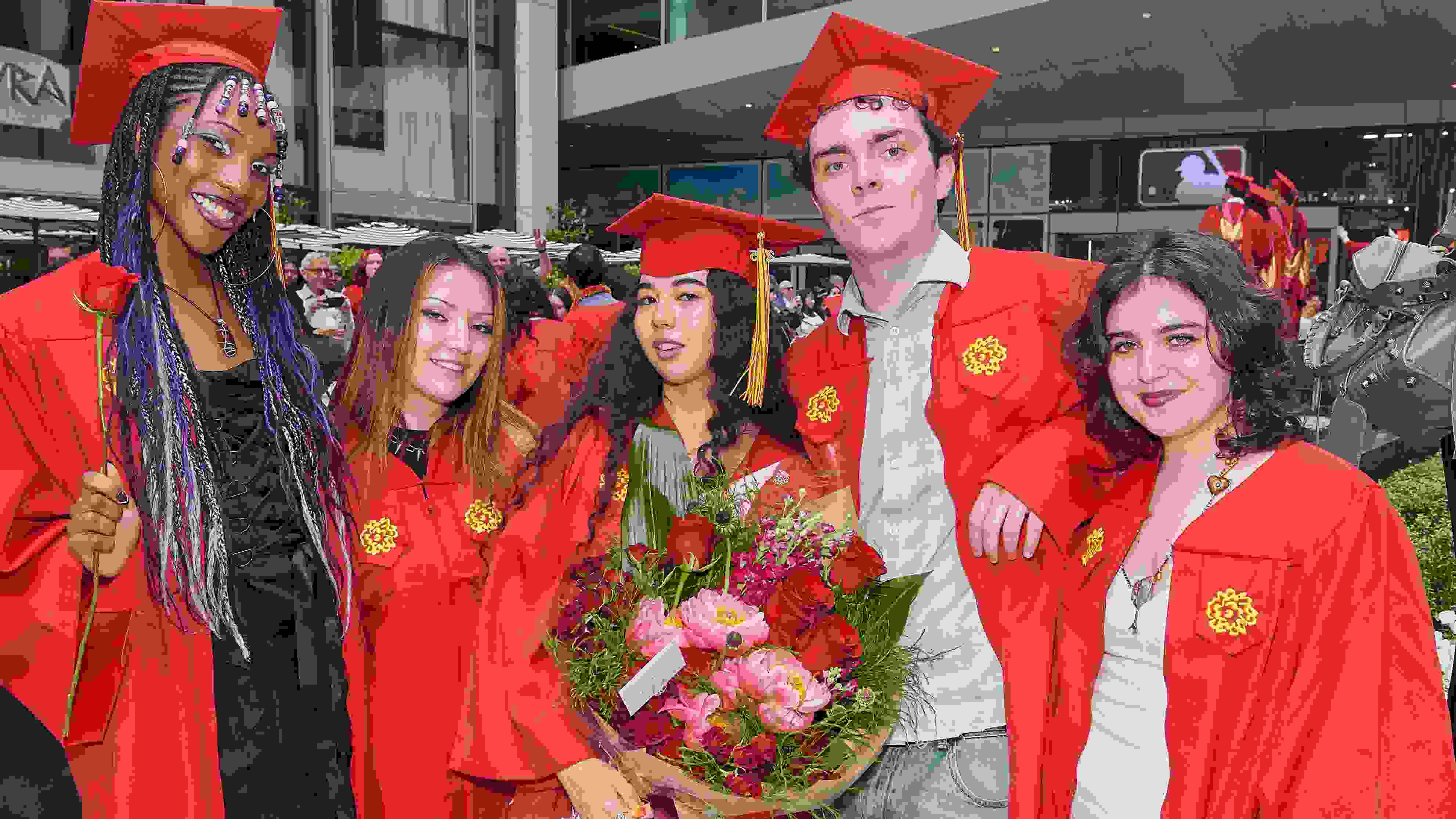 Five graduates wearing red commencement regalia pose for a photo; the middle person is holding a large bouquet of pink and red flowers.