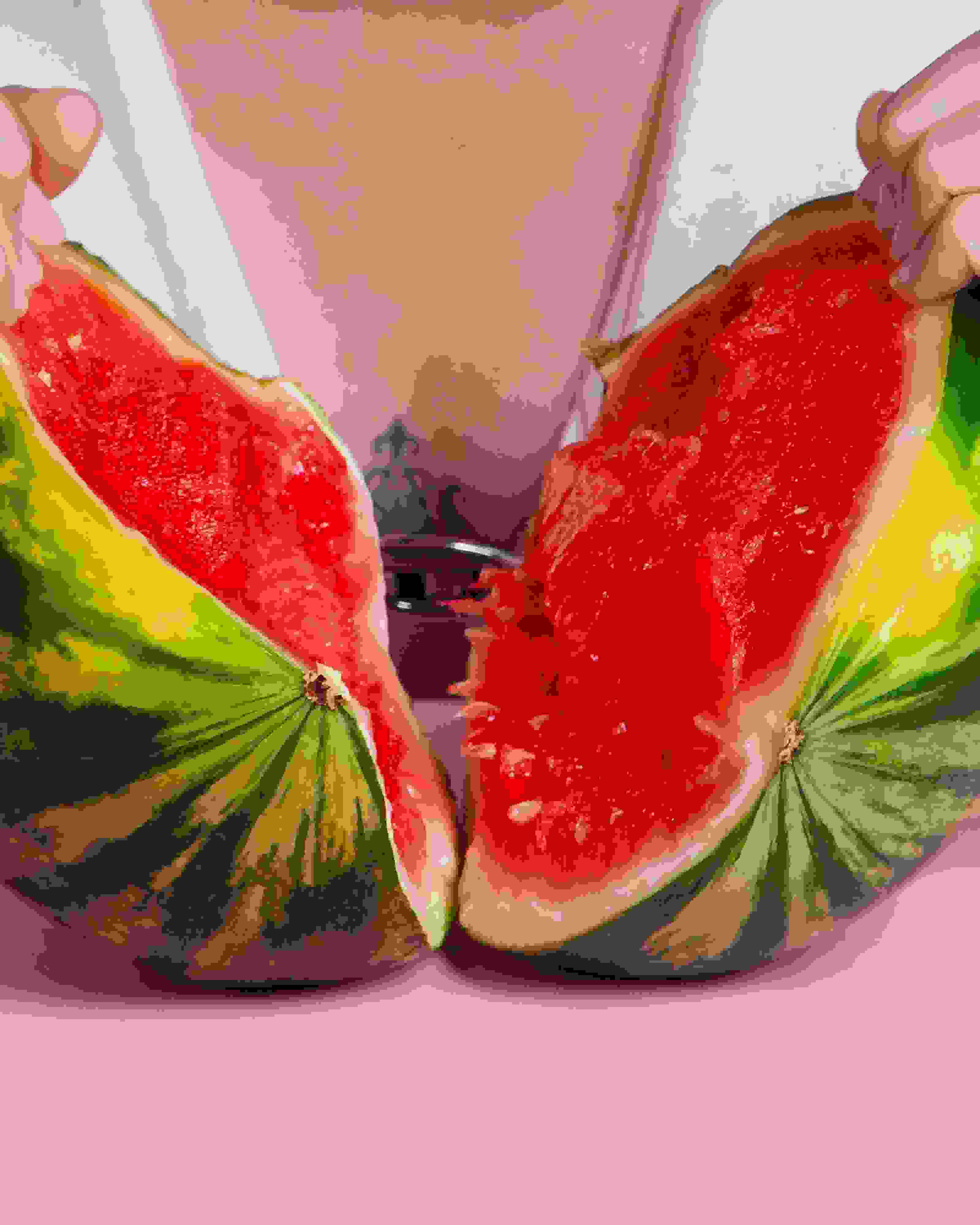 Two hards ripping open a small watermelon in front of a person's chest. There's a pink wall and table in the image as well.