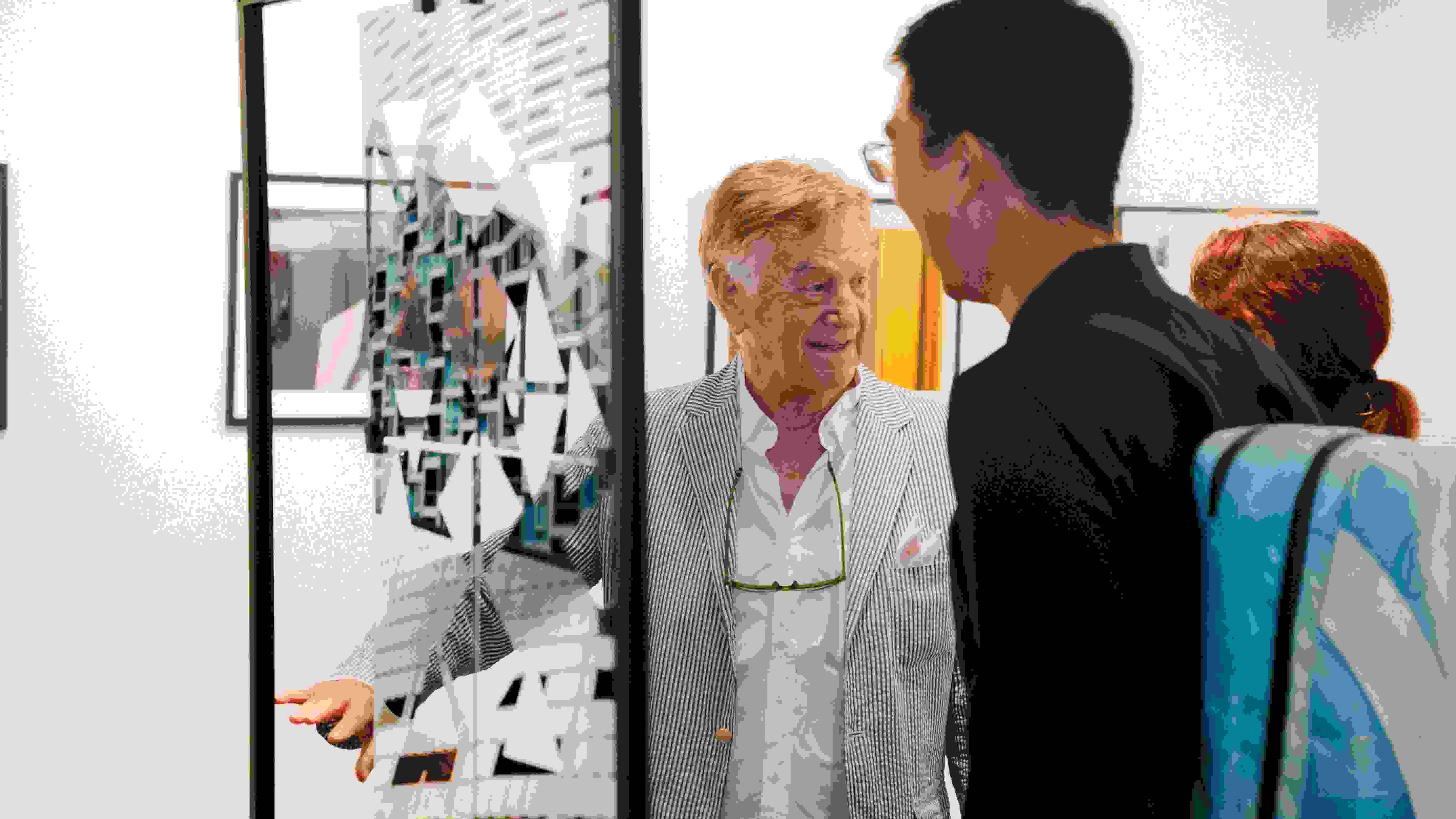 Person wearing a striped blue blazer standing in front of a piece of art talking with two other people.