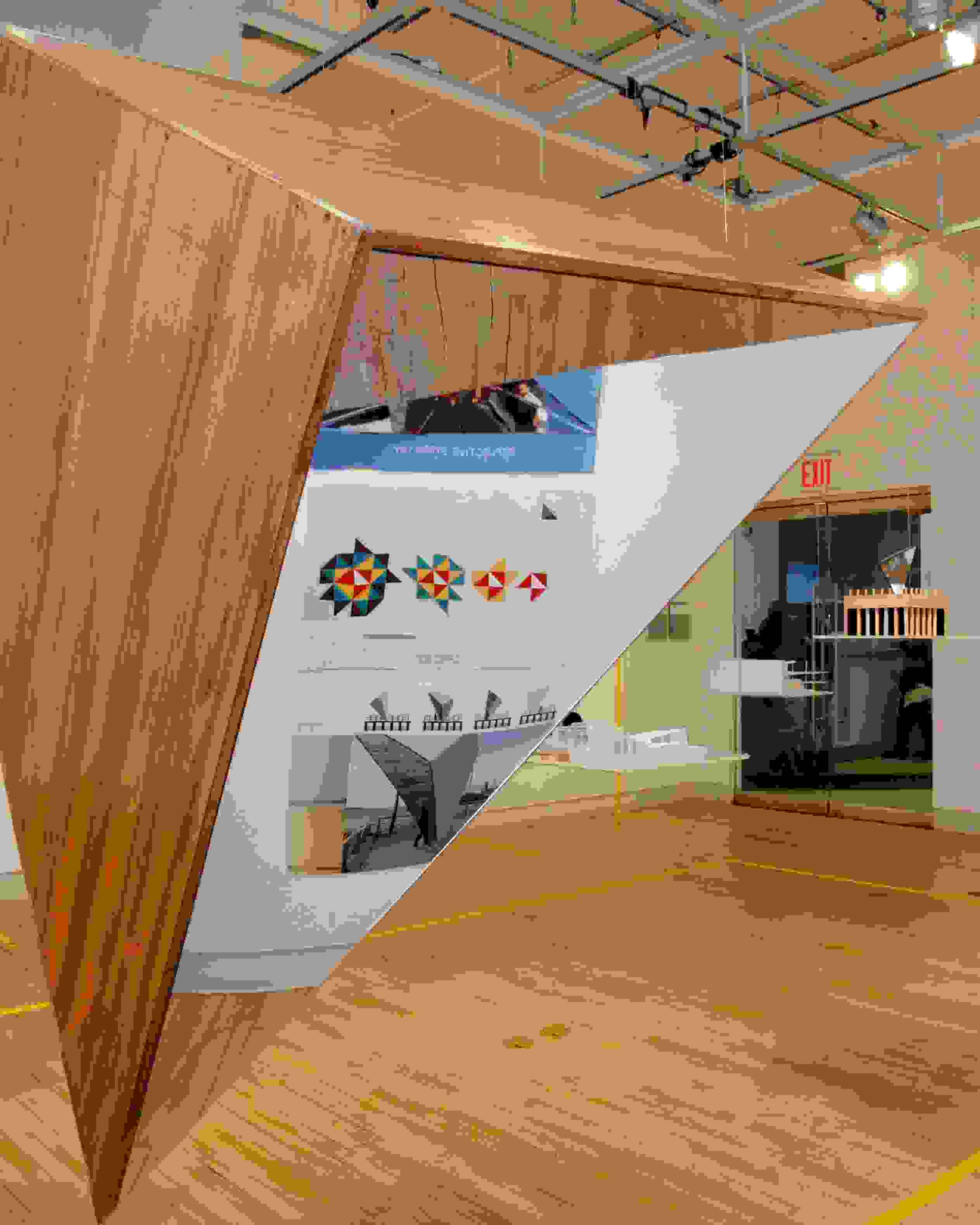 Mirrored pyramid sculpture hangs from the ceiling in the center of the gallery. There are prints on the wall behind the sculpture. On the wall to the right of the sculpture reads, "What's Inside?"