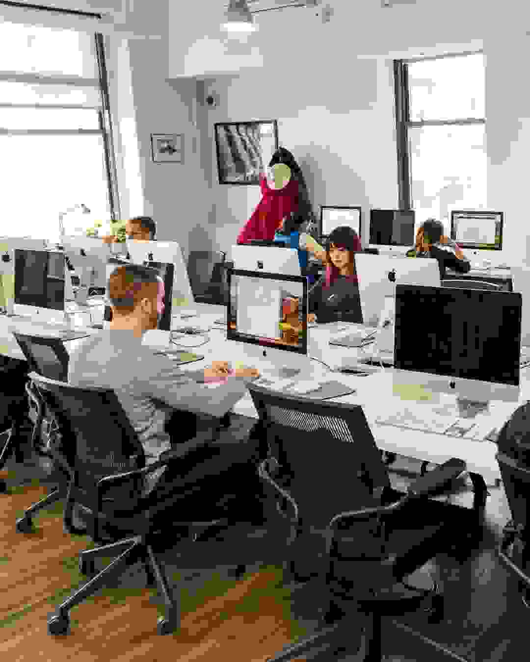 A long table in a computer lab with students sat in office chairs at various Mac desktop computers.