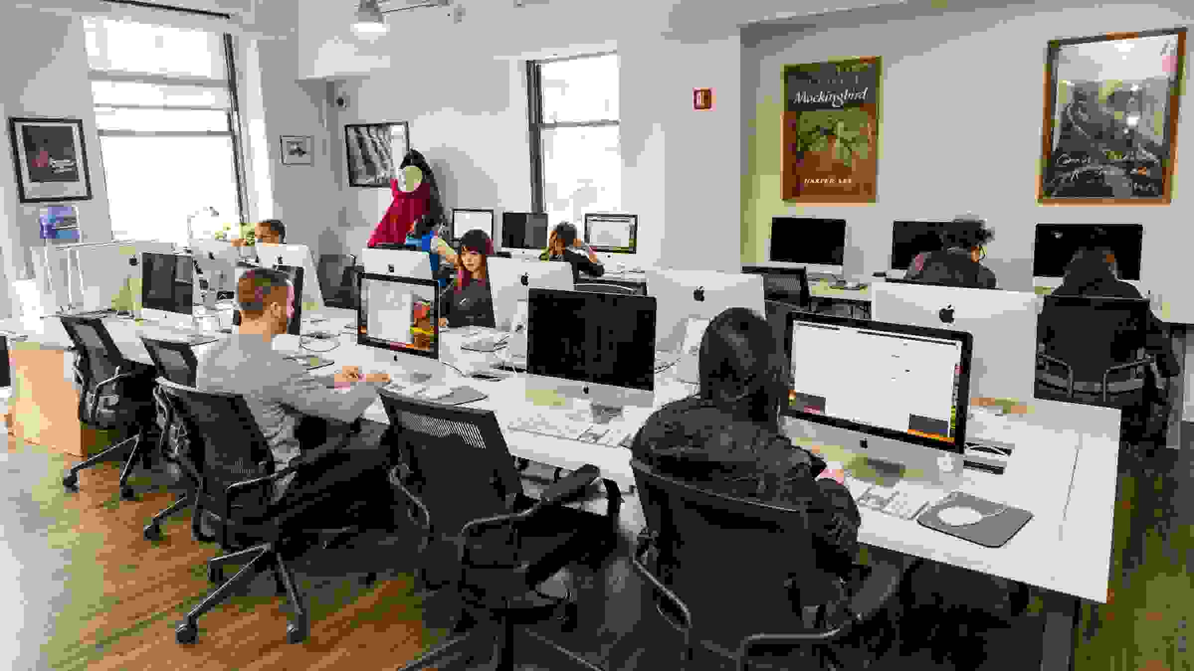 A long table in a computer lab with students sat in office chairs at various Mac desktop computers.