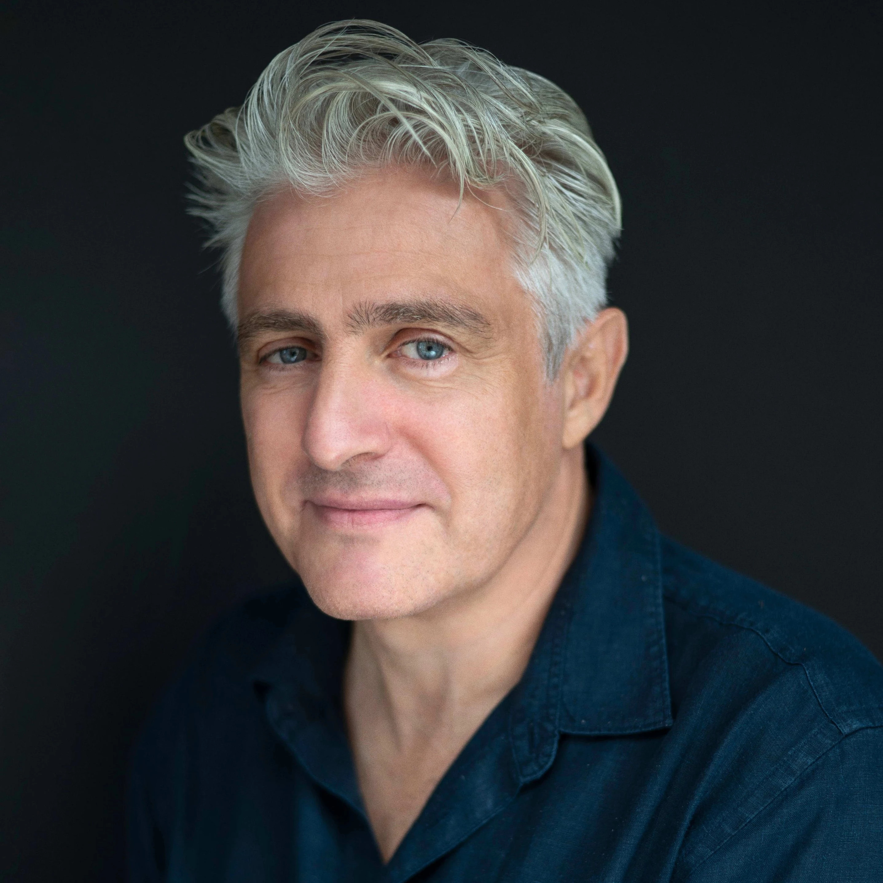 A middle-aged man with grey/white hair in a dark blue shirt looking at the camera upon a black background.