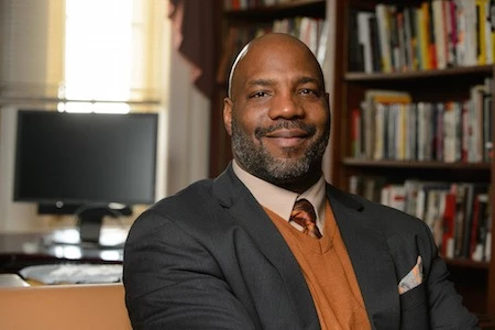 A portrait photograph of writer and academic Jelani Cobb sitting in an office with a desk and bookcase behind him.
