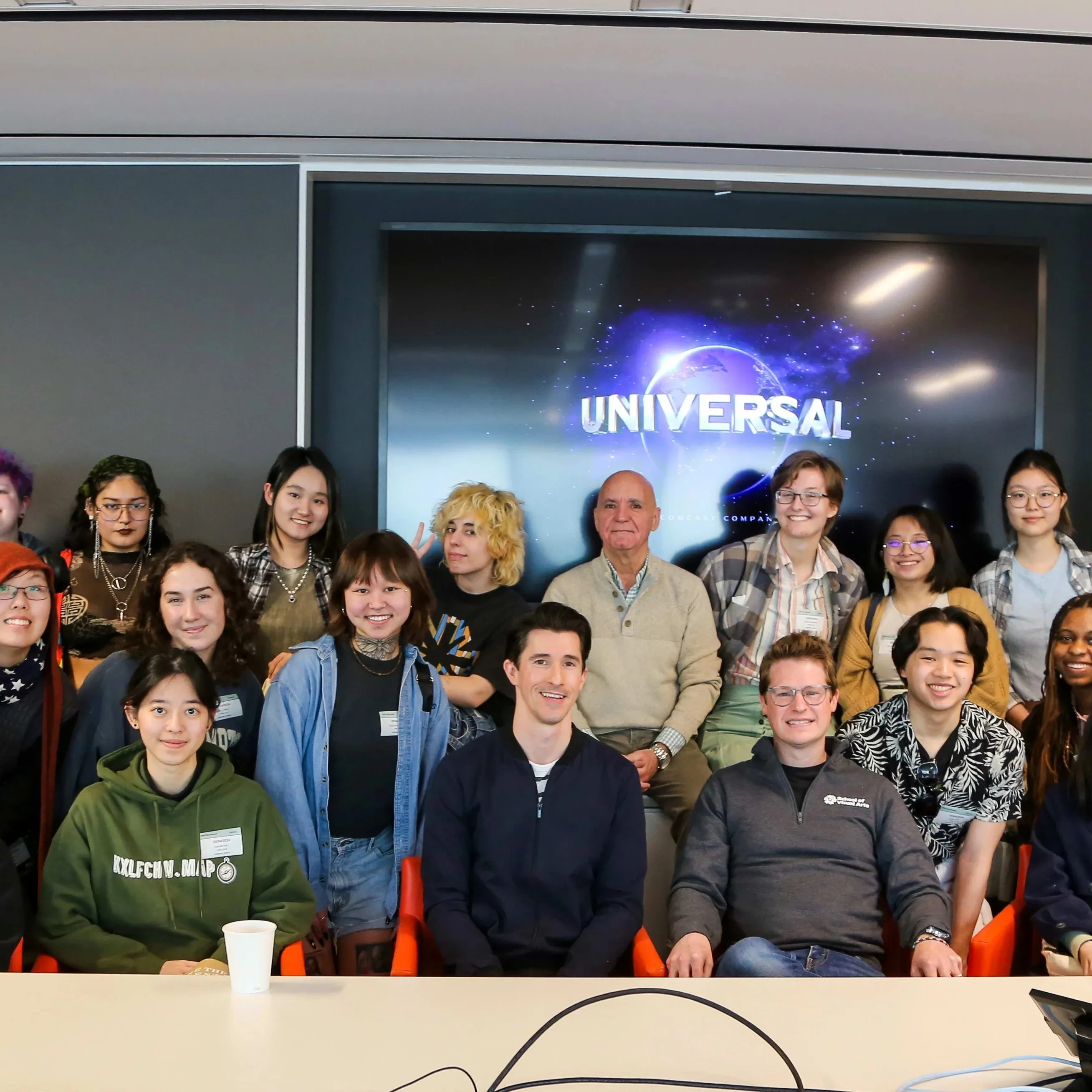 A group of students and adults, likely artists, organized in three rows and posing in front of a screen that displays Universal Studio's name and logo.