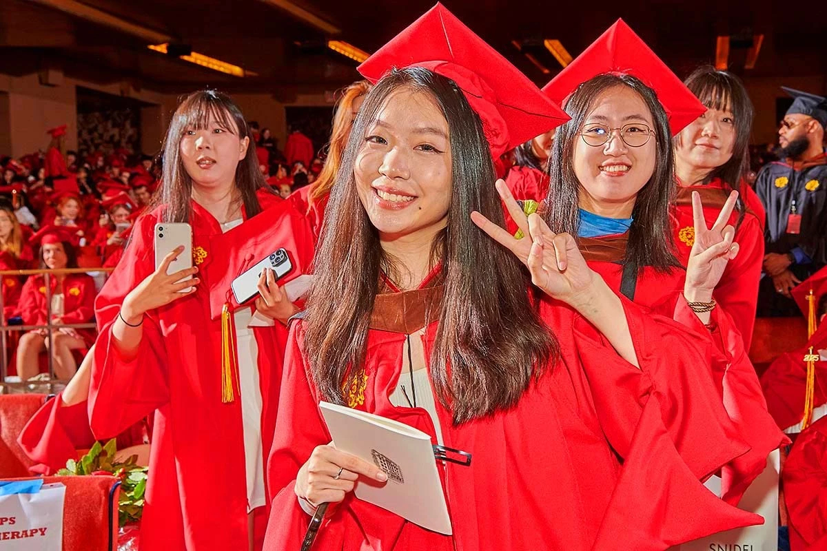 A group of students wearing red graduation gowns and caps smile and hold up peace signs during a graduation ceremony.