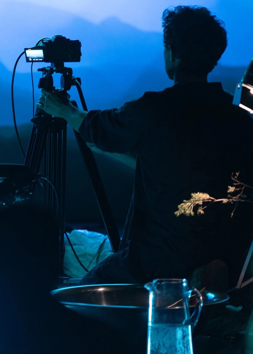  Image of a person pointing a recording camera on a tripod towards a wall with a projected image of what looks like a mountain range.