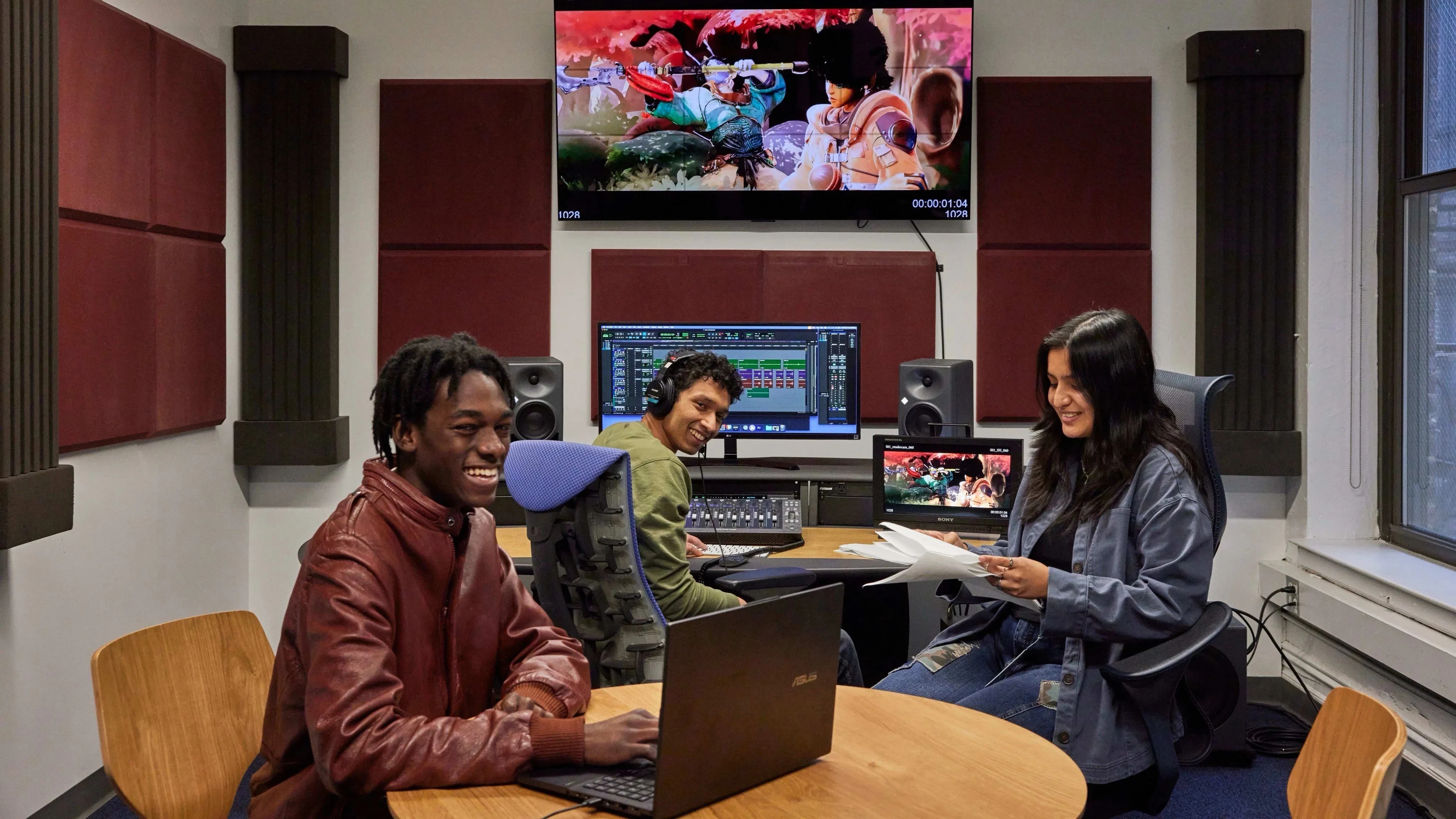 Three students working together in an editing suite with several computers, a large monitor and large speakers.