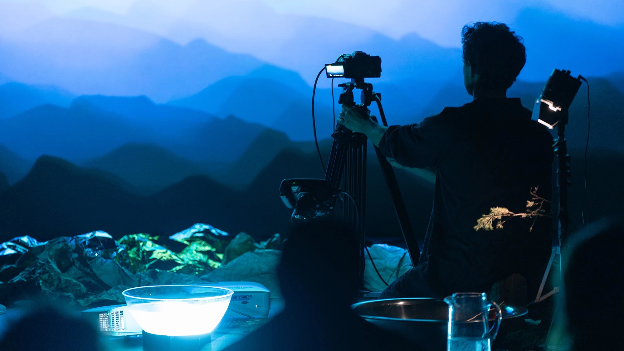  Image of a person pointing a recording camera on a tripod towards a wall with a projected image of what looks like a mountain range.