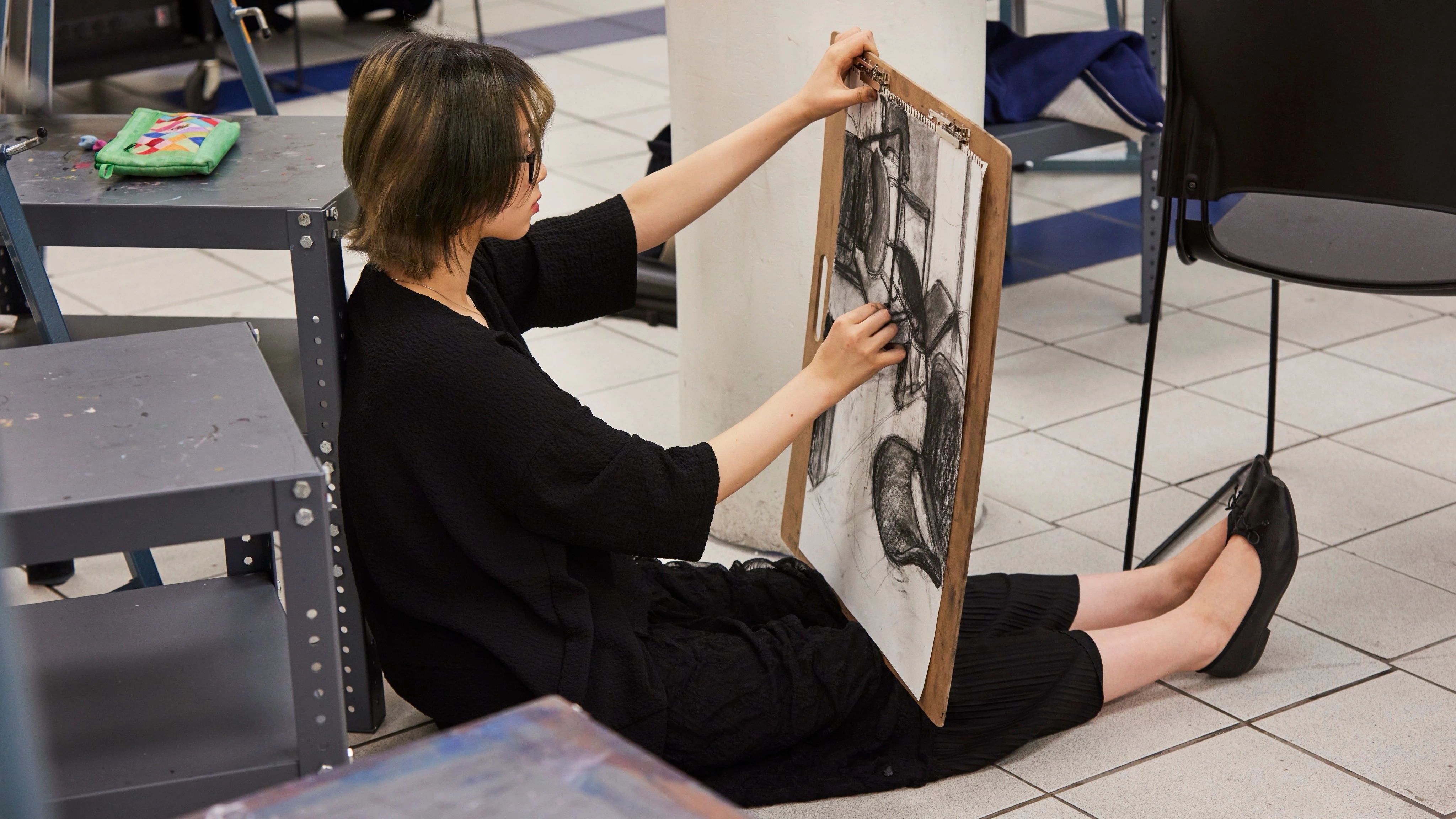 Student sitting on the floor of an art studio sketching a still life with charcoal.