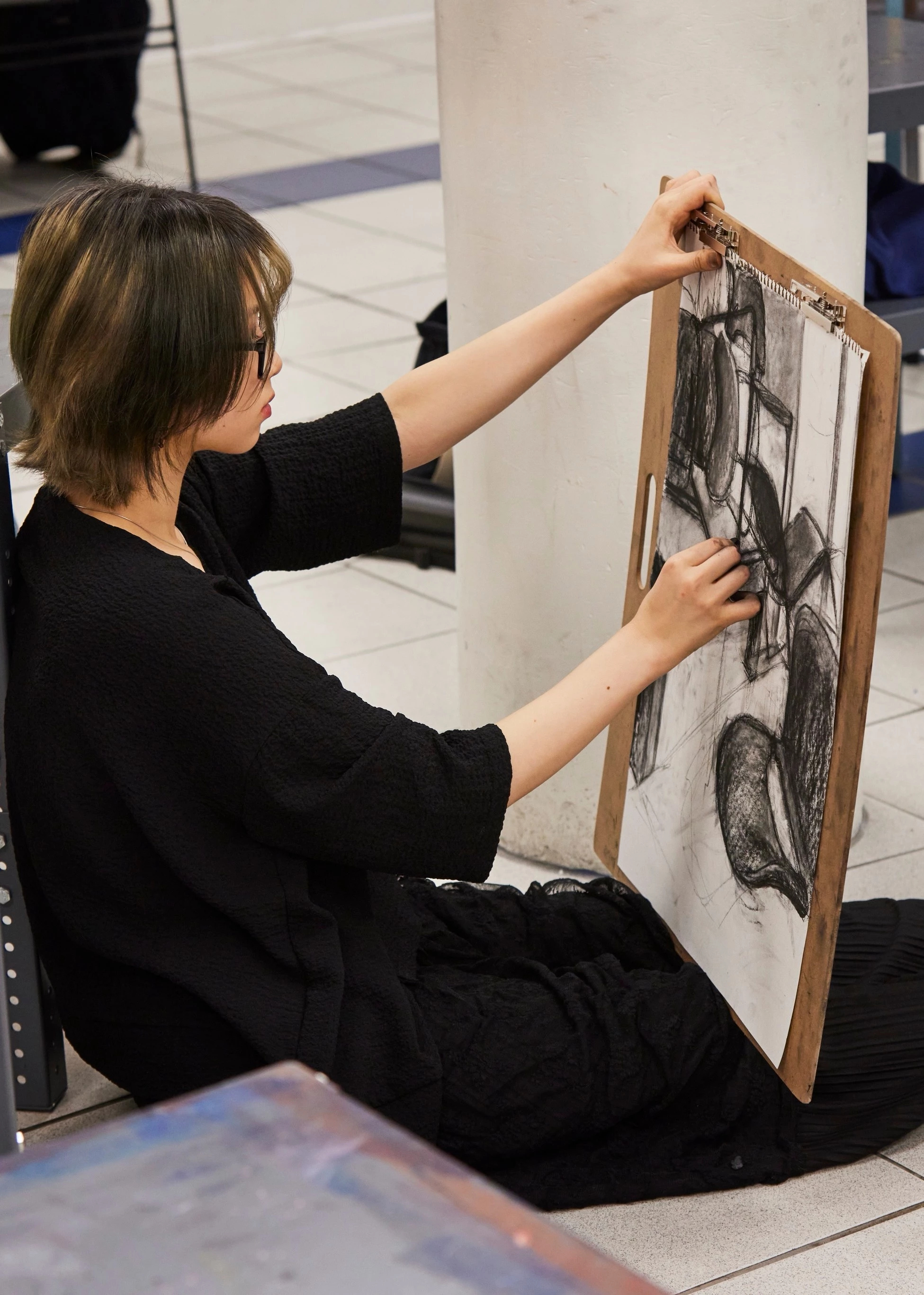 Student sitting on the floor of an art studio sketching a still life with charcoal.