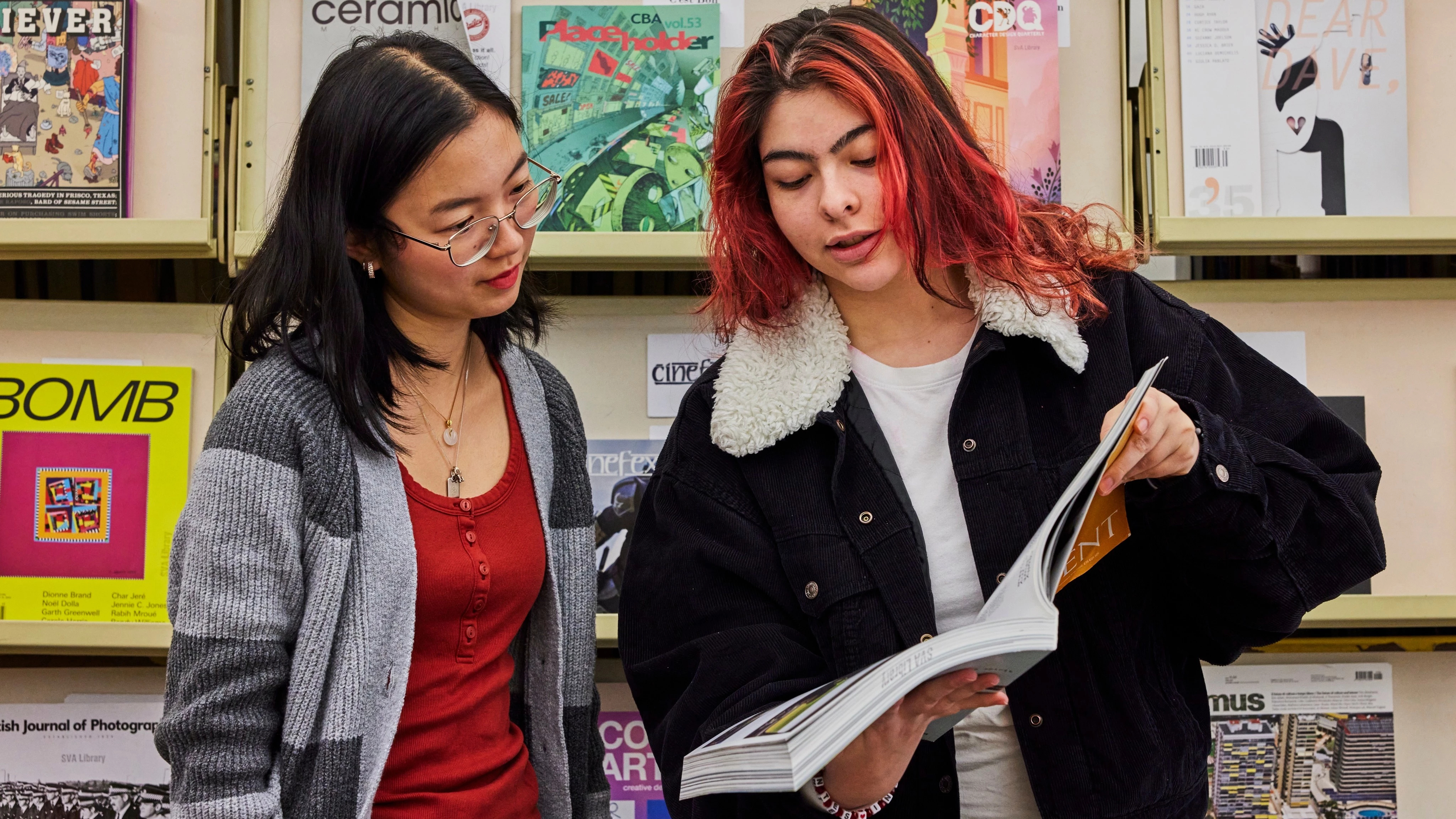 Two students standing up against library stacks reviewing a glossy magazine.