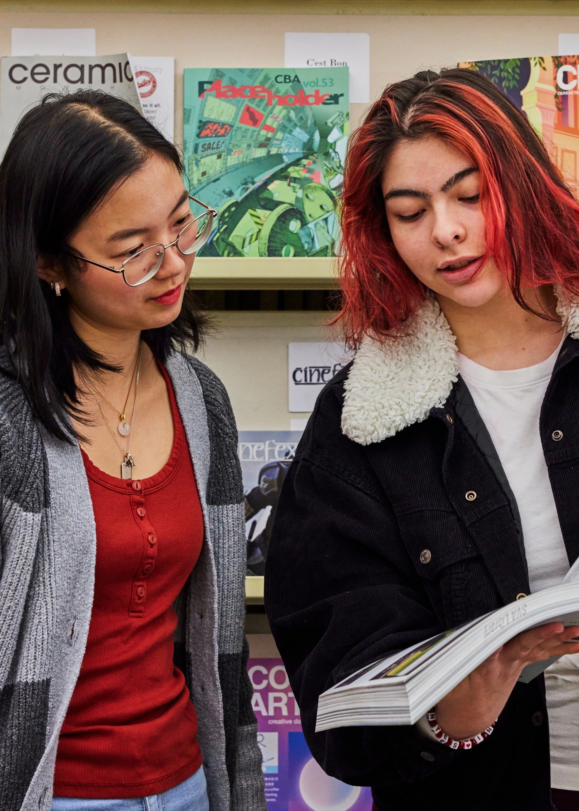 Two students standing up against library stacks reviewing a glossy magazine.