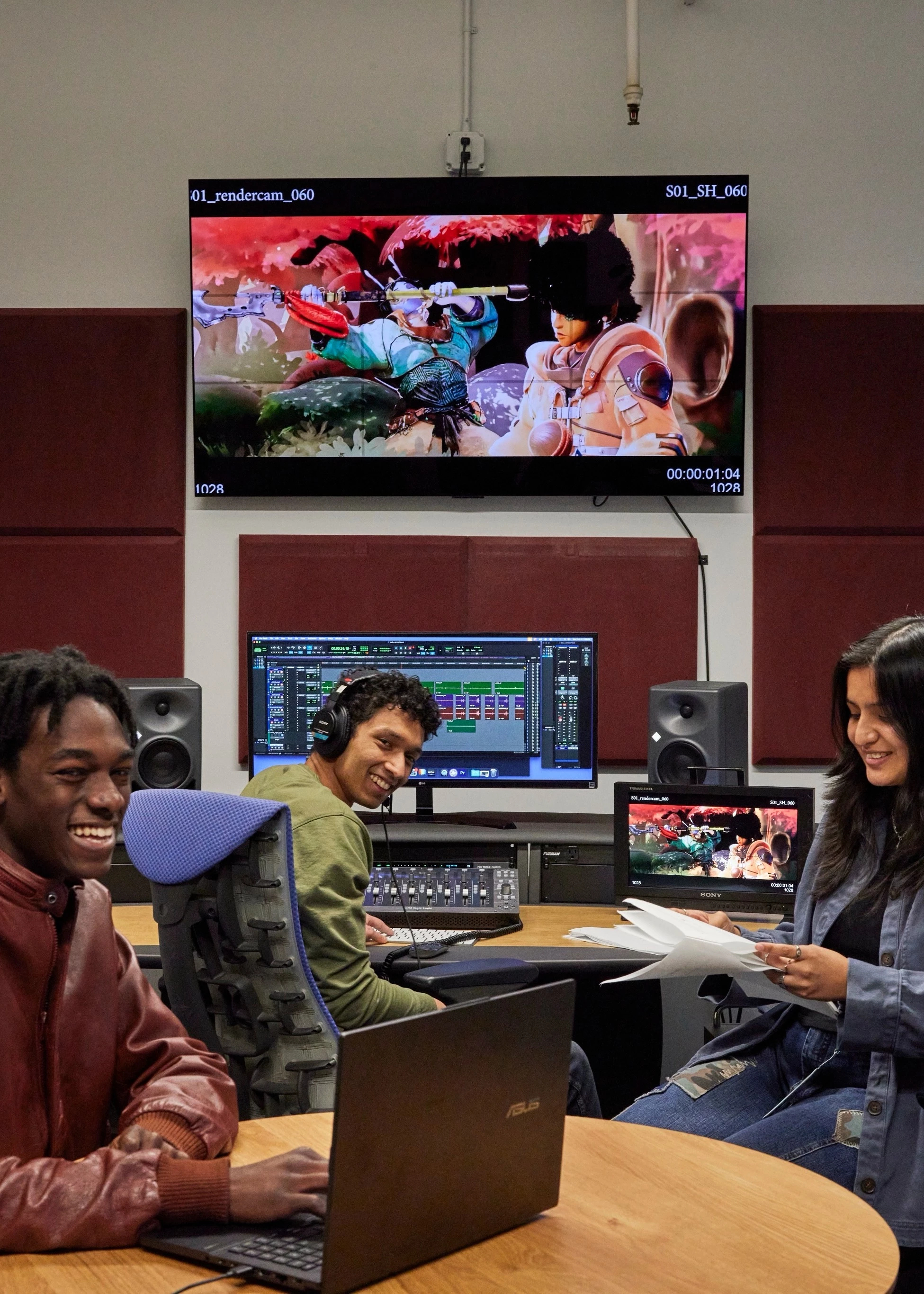 Three students working together in an editing suite with several computers, a large monitor and large speakers.