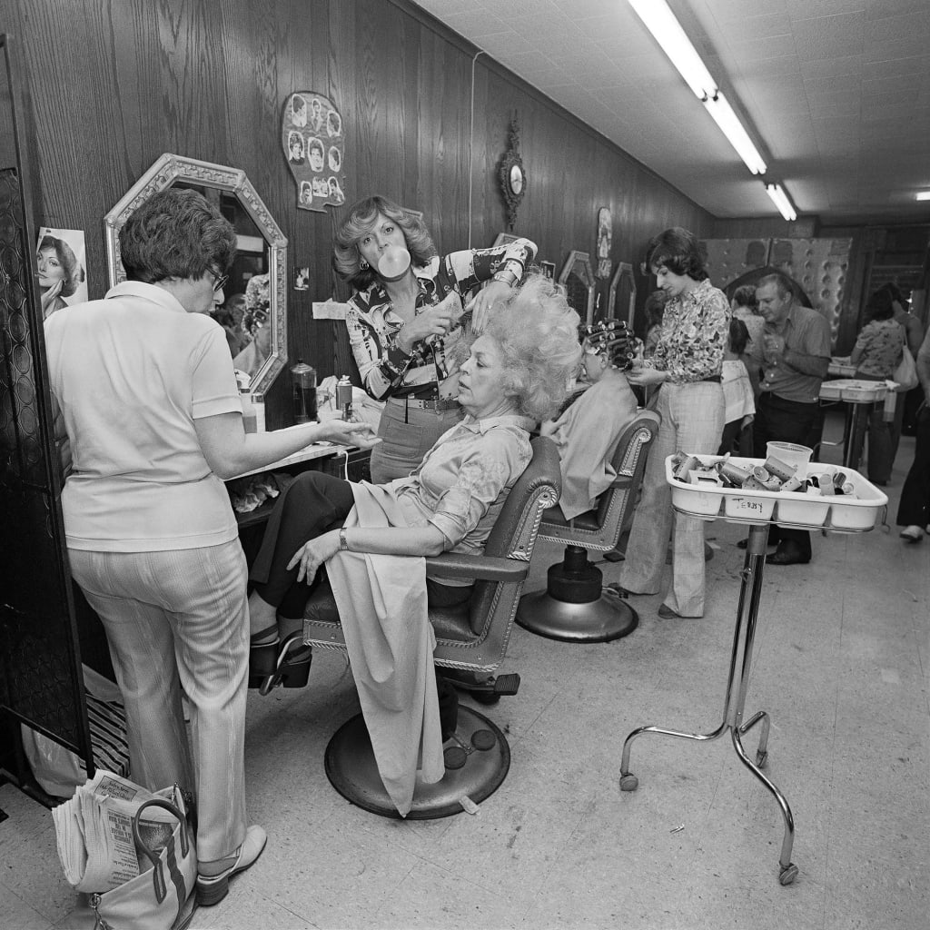 Artist’s mother in a beauty salon getting her hair teased while the beautician blows bubble gum.