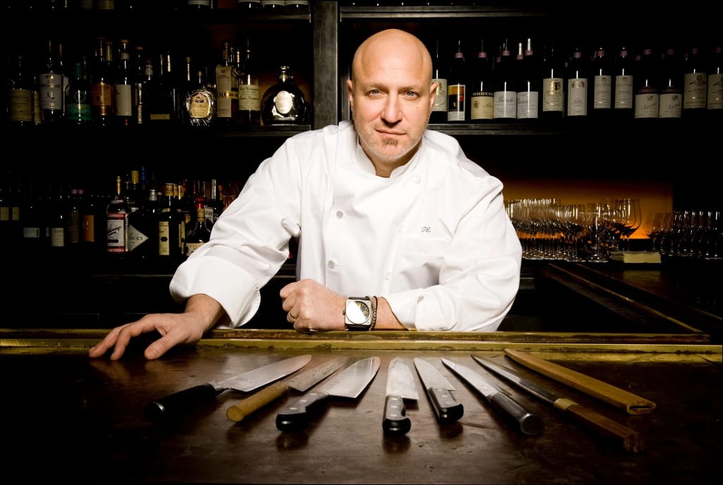 Portrait of Tom Colicchio behind a bar with knives on the counter, all pointed towards him.