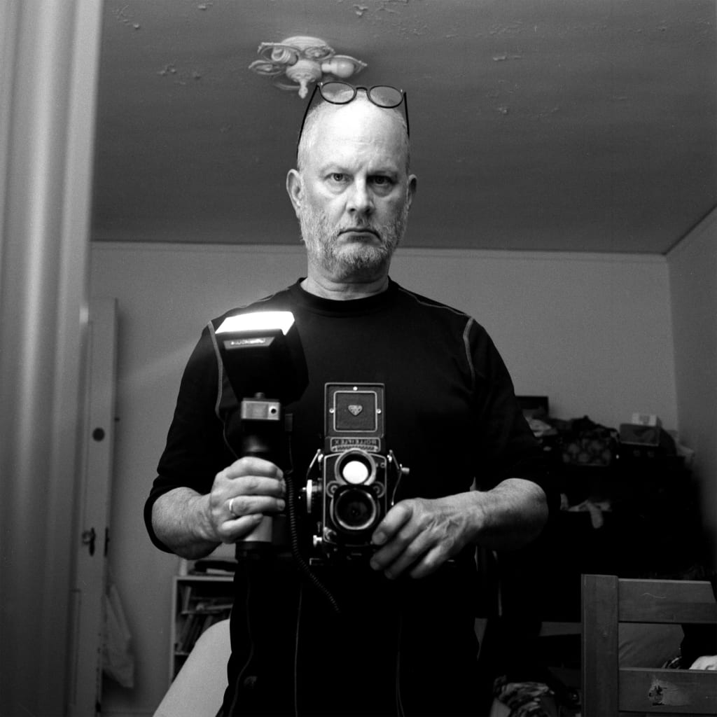 Self-portrait of Jeremiah Dine, an older man. He is wearing a black shirt and is standing in a small room. 