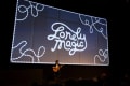 A large slide with the title “ Lonely Magic: is displayed in the background as an individual stands on stage holding an acoustic guitar.