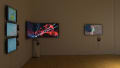 In a gallery space, on the left wall, are three vertically hung monitors playing reels of video art. In the corner are two monitors, one on each wall hung edge-to-edge, playing a video of abstract video work with overlaid text, and to the right is an additional monitor showing a person recording themself from a phone held overhead.