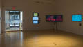 View from inside of gallery space looking toward external glass doors, with three vertically hung monitors to the right of the doors, two hung end-to-end at the corner of the room, and one to its right, each playing video art.