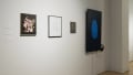Continuing into the other hallway, two framed works are displayed next to a small, dark, unframed work. To the right of them, a monitor displays a large blue fingerprint.