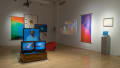 Inside the SVA Flatiron Gallery, colorful banners, monitors, and frames are hung on the walls. A projection is displayed on the right wall, while an installation featuring three televisions and a large, star-like wooden object occupies the center of the room.