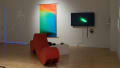 A projection of a woman’s body faintly appears on the left side of another wall. A second abstract print, this time orange, blue, and green, hangs beside her. A monitor is on display to the right of the print, and the red couch sits on the floor in front of it.