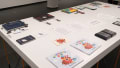 Press releases and a gallery map lay in the center of a table in the gallery. Many different books are arranged around the papers. The nearest book is titled “Bumbling Bud” and depicts a smiling red and orange bumblebee-like creature.