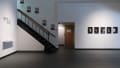 A wall in SVA’s Gramercy Gallery, with a staircase on the left side. Beside it is the name of the exhibition, and to the right are a line of photographs.